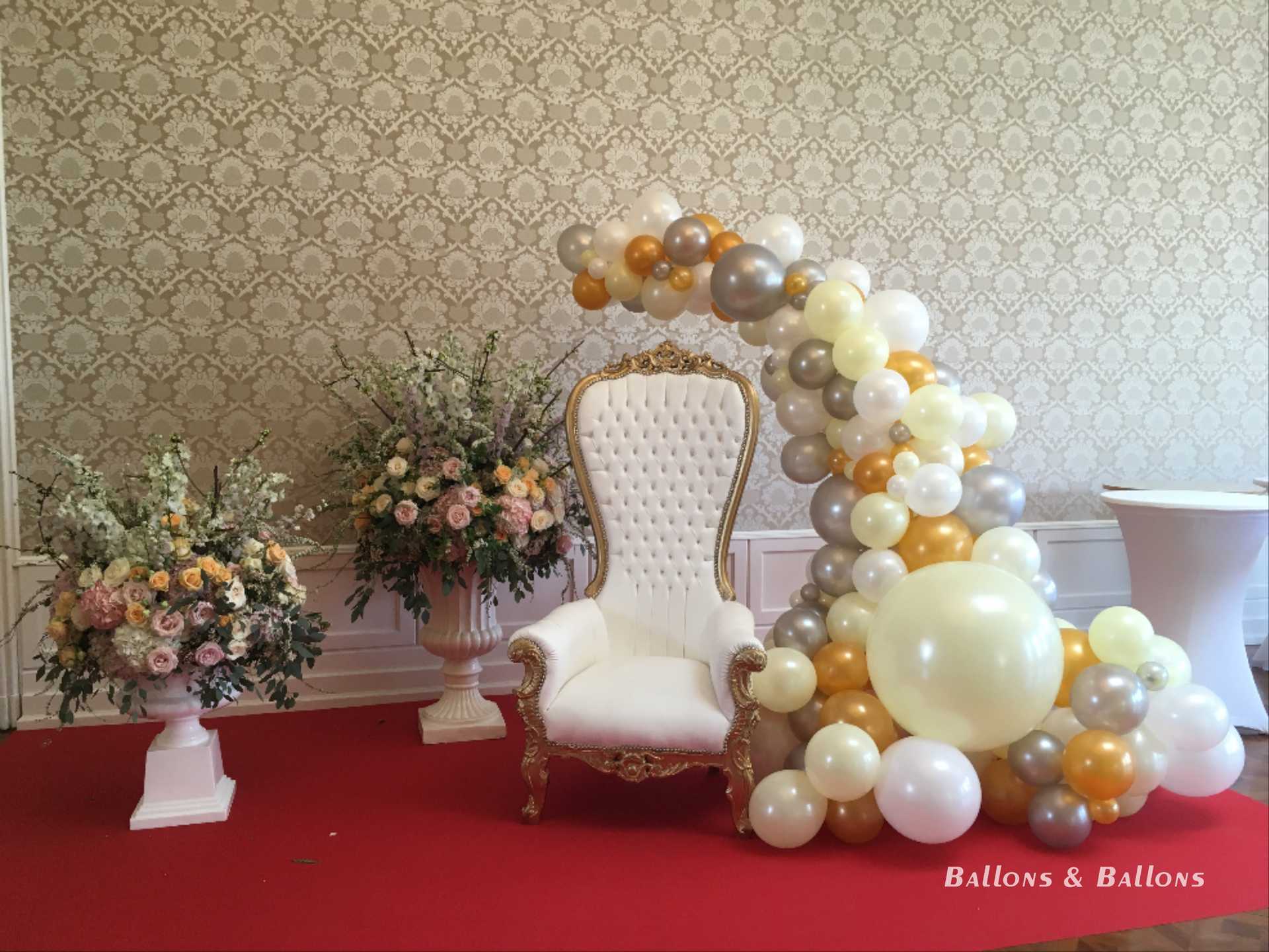 Ein aufwendig dekorierter Raum mit Luftballons, Blumen und einem Stuhl, weißer Stuhl und zwei weiße Vasen mit Blumen.